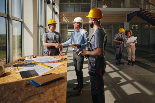 Three men in hard hats discuss commercial construction plans, reviewing building permits and regulations at a planning table.