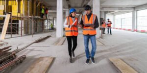 Two men wearing yellow vests in a warehouse, engaged in conversation about the "Building Smarter: Future Design & Build" initiative.