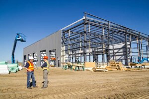 Two men in hard hats stand in front of a large commercial building, representing construction contractors.